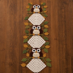 May include: A long, vertical crochet table runner featuring three brown and cream owls with large black and white eyes. The owls are separated by white, diamond-shaped, lacy crochet panels and surrounded by green and yellow crocheted leaves.