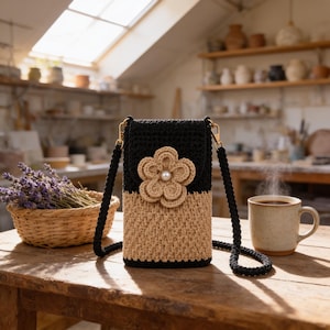 May include: A rectangular crochet crossbody bag featuring a two-tone design with a black upper section and a beige lower section. A large, layered beige crochet flower with a central white pearl accent is attached to the front flap.