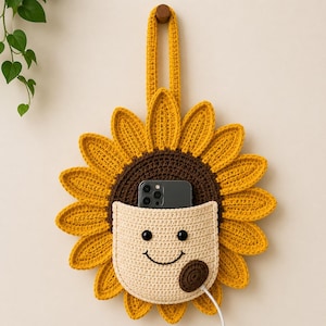 Puede incluir: Un organizador de pared de girasol tejido a mano con un bolsillo para un teléfono. El girasol tiene pétalos amarillos, un centro marrón y un bolsillo beige con una cara sonriente. Una clavija de madera y una correa amarilla están en la parte superior para colgar.