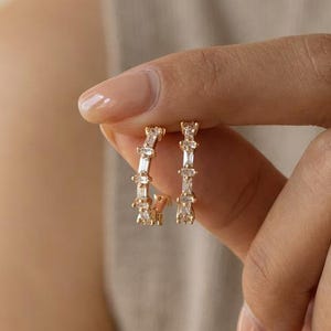 Könnte beinhalten: Ein kleiner goldener Creolen-Ohrring mit einer Reihe klarer, rechteckiger Baguette-Schliff-Steine am äußeren Rand. Das Metall hat eine polierte Oberfläche und die Steine sind in einer durchgehenden Reihe angeordnet, um ein funkelndes, lineares Design zu erzeugen.