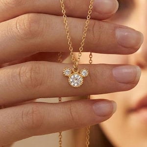 May include: A gold-colored necklace featuring a pendant in the shape of a mouse head. The pendant is adorned with clear, sparkling gemstones. The necklace is held up by a person's hand, showcasing the delicate chain and the pendant's intricate design.