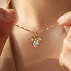 May include: A gold-colored necklace with a pendant shaped like a mouse head. The pendant features a large, round, clear gemstone in the center, with two smaller gemstones on either side. The necklace is being held up by a person.