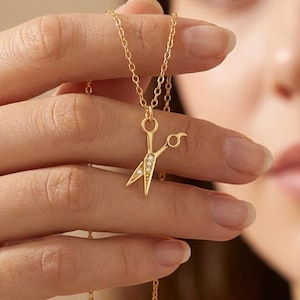 May include: A gold-colored necklace featuring a scissors pendant with small, sparkling embellishments. The scissors design is detailed and hangs from a delicate chain. The necklace is shown against a soft, neutral background.