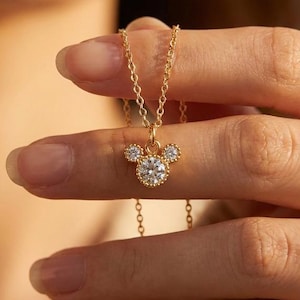 May include: A gold-colored necklace featuring a pendant shaped like a mouse head with three clear, sparkling stones. The necklace is held up by a person's hand, showcasing the delicate chain and pendant design.