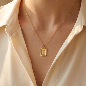 Op de afbeelding: Een goudkleurige ketting met een rechthoekige hanger met de letter "C". De hanger hangt aan een fijne ketting. De persoon draagt een crèmekleurig overhemd met knopen.