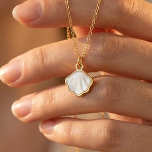 Peut inclure: Un collier doré avec un pendentif en forme de coquillage blanc. Le pendentif est bordé d'or, et la chaîne est fine. L'image présente le collier sur un fond flou.