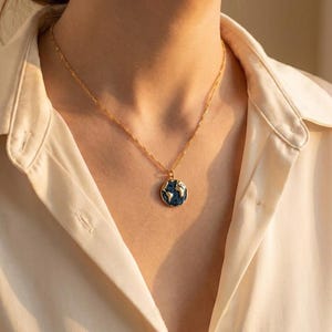 May include: A gold chain necklace featuring a circular pendant with a blue enamel surface and a raised gold map of the world. The pendant hangs from a delicate gold link chain that rests against the collarbone, showcasing a detailed globe design.