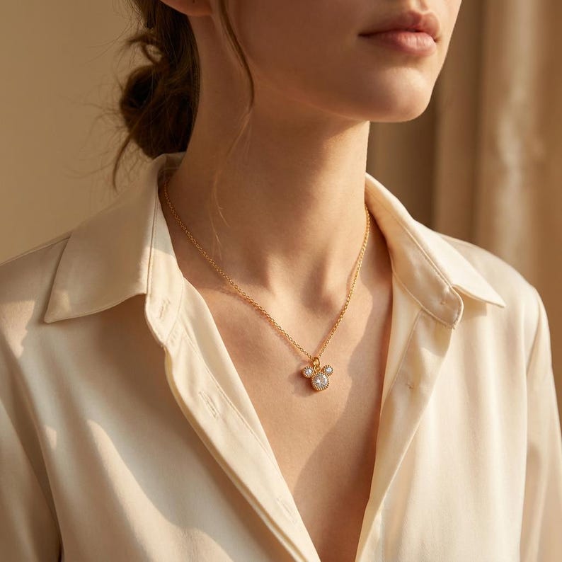 May include: A gold-colored necklace with a pendant featuring a three-stone design. The pendant has a central, larger stone surrounded by smaller stones. The necklace is worn against a cream-colored collared shirt.