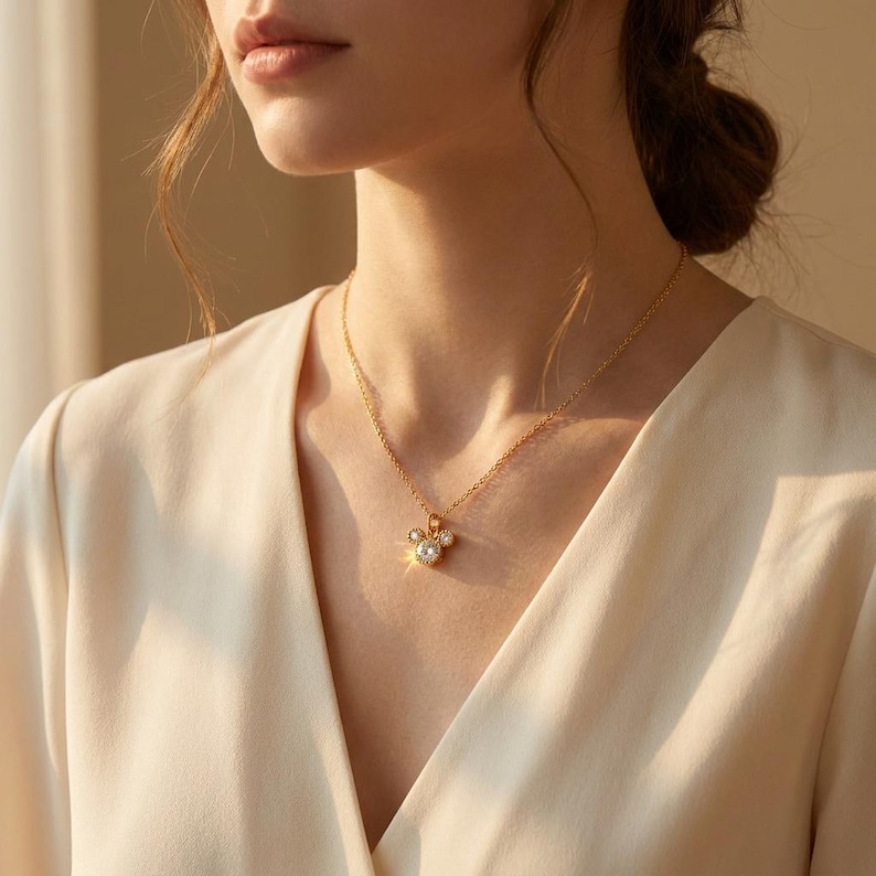 May include: A gold-colored necklace with a pendant featuring a three-circle design, with a central pearl and two smaller pearls. The necklace is worn against a cream-colored top.