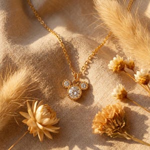 May include: A gold-colored necklace with a pendant shaped like a mouse head, featuring a large, round, clear gemstone in the center and smaller gemstones for the ears. The necklace is on a beige fabric background, with dried flowers.