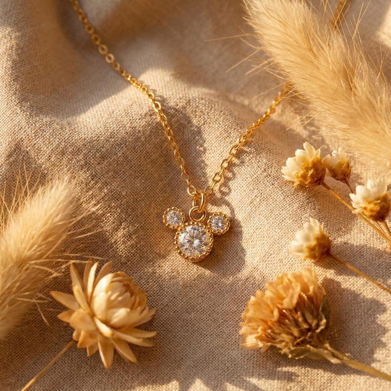 May include: A gold-colored necklace with a pendant shaped like a mouse head, featuring a large, round, clear gemstone in the center and smaller gemstones for the ears. The necklace is on a beige fabric background, with dried flowers.