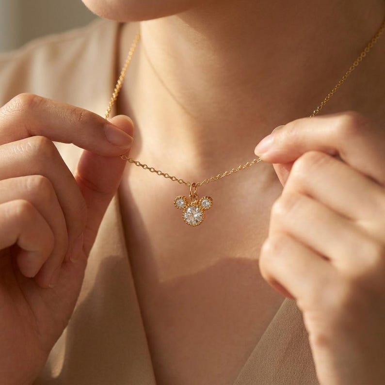 May include: A gold-colored necklace with a pendant shaped like a mouse head. The pendant features a large, round, clear gemstone in the center, with two smaller gemstones on either side. The necklace is being held up by a person.