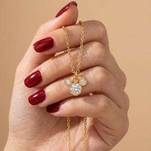 May include: A gold-colored necklace featuring a pendant in the shape of a mouse head. The pendant has a large, round, clear gemstone for the face and two smaller gemstones for the ears. The necklace is held up by a hand with red nail polish.