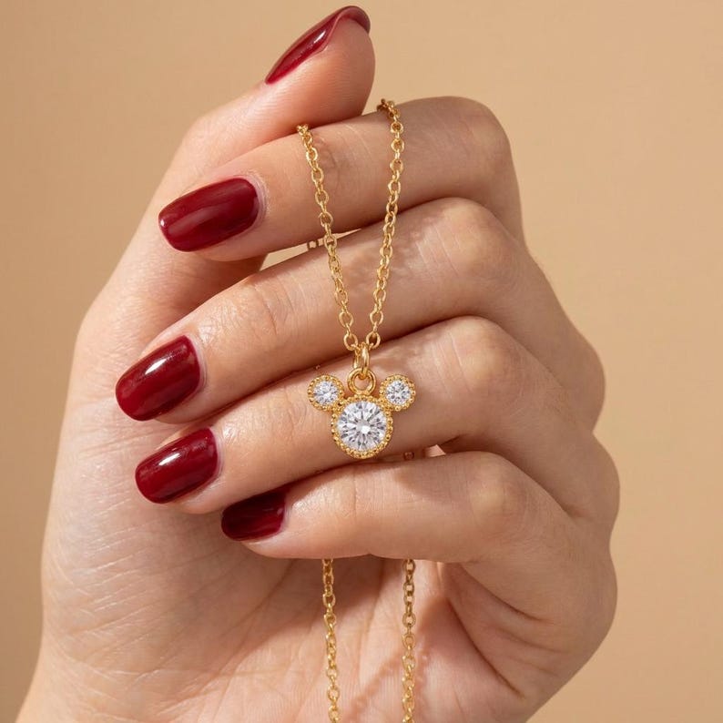 May include: A gold-colored necklace featuring a pendant in the shape of a mouse head. The pendant has a large, round, clear gemstone for the face and two smaller gemstones for the ears. The necklace is held up by a hand with red nail polish.
