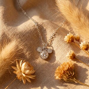 May include: A silver necklace with a pendant shaped like a mouse head, featuring a large, round, sparkling center stone and two smaller side stones. The necklace is displayed on a beige fabric with dried flowers.