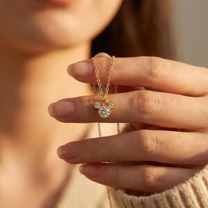 May include: A gold-colored necklace featuring a pendant shaped like a mouse head with three clear, sparkling stones. The necklace is held up by a person's hand, showcasing the delicate chain and pendant design.