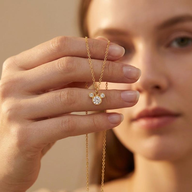 May include: A gold-colored necklace featuring a pendant in the shape of a mouse head. The pendant is adorned with clear, sparkling gemstones. The necklace is held up by a person's hand, showcasing the delicate chain and the pendant's intricate design.