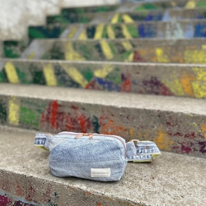 Könnte beinhalten: Hellblaue Jeans-Bauchtasche mit kupferfarbenem Reißverschluss und einem kleinen weißen Etikett mit der Aufschrift "Less new. More stuff." Die Tasche liegt auf einer bemalten Betonstufe.