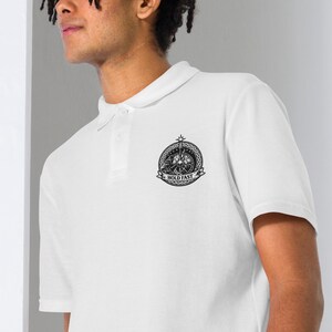 May include: White polo shirt with a black and white emblem on the left chest. The emblem features a circular design with the words "HOLD FAST" on a banner. The shirt has a classic collar and button placket.