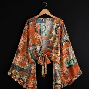 May include: An orange kimono-style top with a patchwork design in shades of orange, brown, and blue. The top has long, wide sleeves and a tie closure at the waist. The garment is hanging on a wooden hanger against a black background.