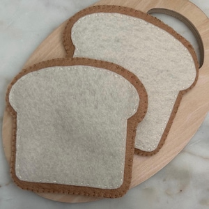 May include: Two white felt pieces shaped like slices of bread on a wooden cutting board.