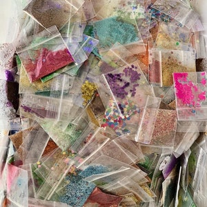 May include: A large collection of small plastic bags filled with various colors of glitter and sequins. The bags are piled on top of each other in a random order.