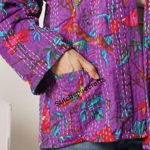 May include: A purple quilted jacket featuring a vibrant floral and bird pattern in shades of red, pink, and teal. The garment is detailed with white running stitch embroidery throughout and includes a front pocket with the text Stitchspirecrafts.