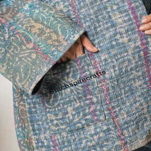 May include: A blue quilted jacket featuring a faded floral pattern in shades of teal, grey, and white. The garment is detailed with visible pink running stitch embroidery throughout and includes a front patch pocket with the text Stitchspirecrafts.