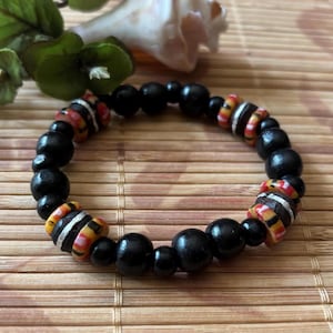 May include: A beaded bracelet featuring black wooden beads interspersed with colorful, patterned beads in red, orange, yellow, and white. The bracelet is displayed on a natural, textured surface.