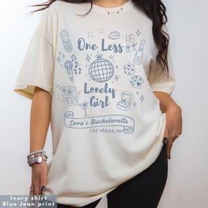 Puede incluir: Camiseta de color marfil con estampado azul vaquero. La camiseta presenta el texto "One Less Lonely Girl" con ilustraciones de una bola de discoteca, champán y un joyero. El texto "Lora's Bachelorette Las Vegas, NV" también está impreso.