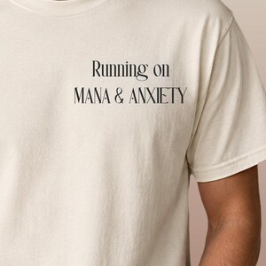 Puede incluir: Una camiseta de manga corta color crema con texto negro impreso en la parte superior izquierda del pecho. El texto dice Running on MANA & ANXIETY en una fuente serif estilizada, con pequeños símbolos de estrellas sobre las letras n y A.