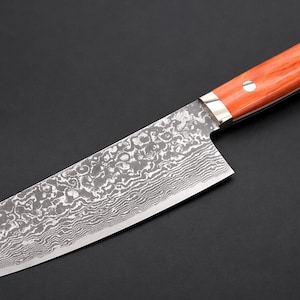 以下が含まれることがあります： 刃に模様が入ったステンレス製のシェフナイフと木製の柄。 刃は独特の質感のあるデザインです。 柄は赤茶色で、2つの銀色のリベットで固定されています。 ナイフは黒い背景に配置されています。