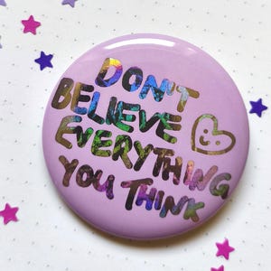 Könnte beinhalten: Ein runder, lavendelfarbener Ansteckbutton mit dem irisierenden Text "DON'T BELIEVE EVERYTHING YOU THINK" und einem kleinen Herzsymbol mit einem lächelnden Gesicht. Die Schrift hat ein mehrfarbiges, reflektierendes Finish.