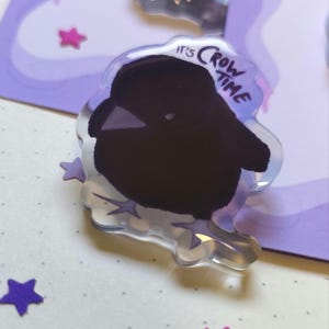 Könnte beinhalten: Ein klarer Acryl-Pin mit einer schwarzen Krähenillustration und dem Text "It's Crow Time". Der Pin ist von lila sternförmigem Konfetti und einem lila Hintergrund umgeben.