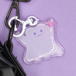 Può includere: Un portachiavi a forma di fantasma in acrilico trasparente con un volto sorridente e un fiocco rosa. Il fantasma è riempito di glitter e ha una chiusura bianca attaccata a un moschettone nero. Lo sfondo è viola.