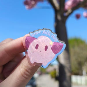 Könnte beinhalten: Eine durchscheinende Acryl-Anstecknadel mit einem hellrosa, runden Wesen, dunkelrosa Ohren und einem kleinen gelben Stern. Die Anstecknadel hat eine glitzernde Oberfläche und den Schriftzug "brave little thing" in hellblauer Schreibschrift.