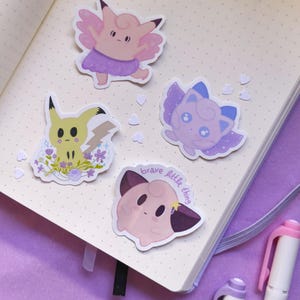 Könnte beinhalten: Vier farbenfrohe Aufkleber mit Zeichentrickfiguren sind auf einem Notizbuch angeordnet. Die Aufkleber zeigen eine rosa Fee, eine gelbe Figur mit einem Blitz und zwei weitere Figuren in Rosa- und Lilatönen. Ein Aufkleber trägt den Text "brave little thing".