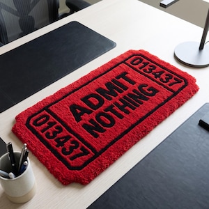 Puede incluir: Una alfombra rectangular roja con mechones en forma de boleto, con texto negro que dice ADMIT NOTHING y el n&uacute;mero 013437 impreso en negro en las esquinas superior e inferior. La alfombra tiene un pelo grueso y texturizado de alto contraste.