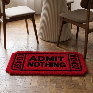 Puede incluir: Una alfombra rectangular roja con forma de boleto vintage con bordes negros y texto negro que dice ADMIT NOTHING. La alfombra presenta el n&uacute;mero 013437 impreso en negro tanto en el lado izquierdo como en el derecho del dise&ntilde;o del boleto.