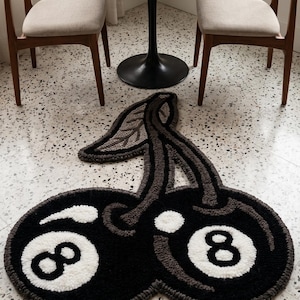 Könnte beinhalten: Ein getufteter Teppich in Form von zwei Kirschen, wobei jede Kirsche wie eine schwarze Acht-Ball-Billardkugel mit einem weißen Kreis und der Zahl Acht gestaltet ist. Der Teppich hat einen dunkelgrauen Stiel und ein Blatt mit dickem Flor.