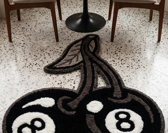 Puede incluir: Una alfombra de mechones con forma de dos cerezas, donde cada cereza está diseñada para parecerse a una bola de billar negra número ocho con un círculo blanco y el número ocho. La alfombra presenta un tallo y una hoja gris oscuro.