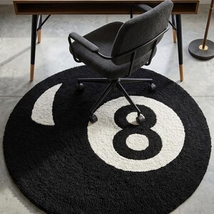 May include: A round black rug with a white number eight design. The rug is placed on a gray floor, with a desk, chair, and window in the background. The rug's design resembles a billiard ball.