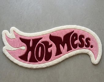 Puede incluir: Una alfombra de mechones con un fondo rosa en forma de llama y un borde blanco. El texto "Hot Mess." está escrito en una fuente estilizada de color granate oscuro en el centro de la alfombra. El diseño tiene una superficie suave y texturizada.