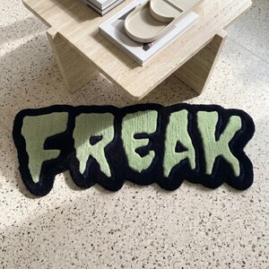May include: A tufted rug featuring the word FREAK in light green letters with a dark navy blue outline. The letters have a dripping, irregular shape and a textured pile surface. The rug is positioned on a light-coloured speckled floor.