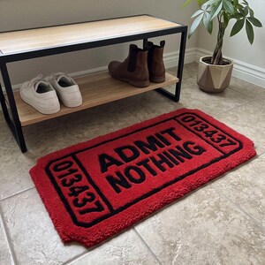 Puede incluir: Una alfombra rectangular roja con superficie texturizada, borde negro y el texto ADMIT NOTHING en letras negras en negrita. La alfombra est&aacute; dise&ntilde;ada para parecerse a un boleto antiguo con el n&uacute;mero 013437 impreso en ambos extremos en color negro.