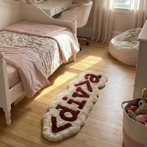 May include: A cream-colored rug with the name "Livia" in a deep red, placed on a hardwood floor. The rug is in a child's bedroom, with a white bed, desk, and chair visible. A basket of toys sits nearby.