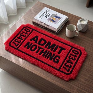 Puede incluir: Una alfombra rectangular roja de tejido tufting con un borde negro y el texto "ADMIT NOTHING" en letras negras negritas. El dise&ntilde;o imita un boleto vintage, con el n&uacute;mero "013437" impreso en negro en los extremos izquierdo y derecho.