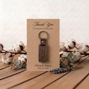 May include: A wooden keychain with a leather strap and metal ring is displayed on a card that reads "Thank You". The keychain is engraved with "Olivia & James" and the date "June 15, 2027". The card is on a wooden surface.
