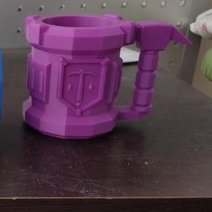 Könnte beinhalten: Drei 3D-gedruckte Becher mit geometrischem, festungsartigem Design, jeweils mit einem Schildemblem und einem hammerförmigen Henkel. Die Becher sind mit Marble, Blue und Purple beschriftet, was ihren jeweiligen Farben Weiß, Blau und Lila entspricht.