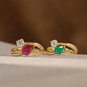 Peut inclure: Deux bagues en or, chacune ornée d'une pierre précieuse blanche étincelante et d'une pierre précieuse en forme de poire colorée. Une bague a une pierre rouge, l'autre une pierre verte. Les bagues sont présentées sur une surface neutre.